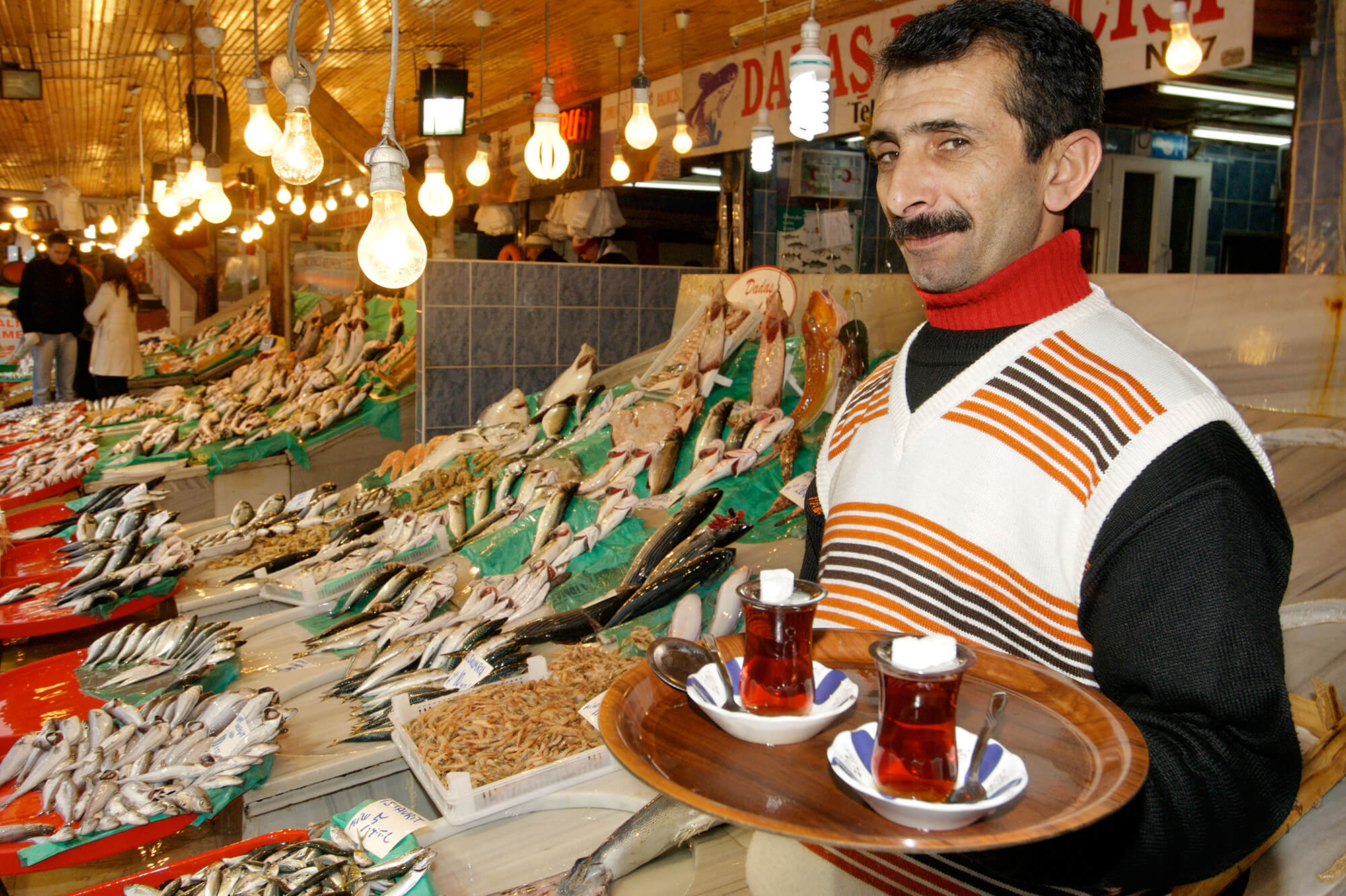 Colourful photograph of Mehmed, the Foodbar Barabas mascot, at a fish market serving tea and smiling