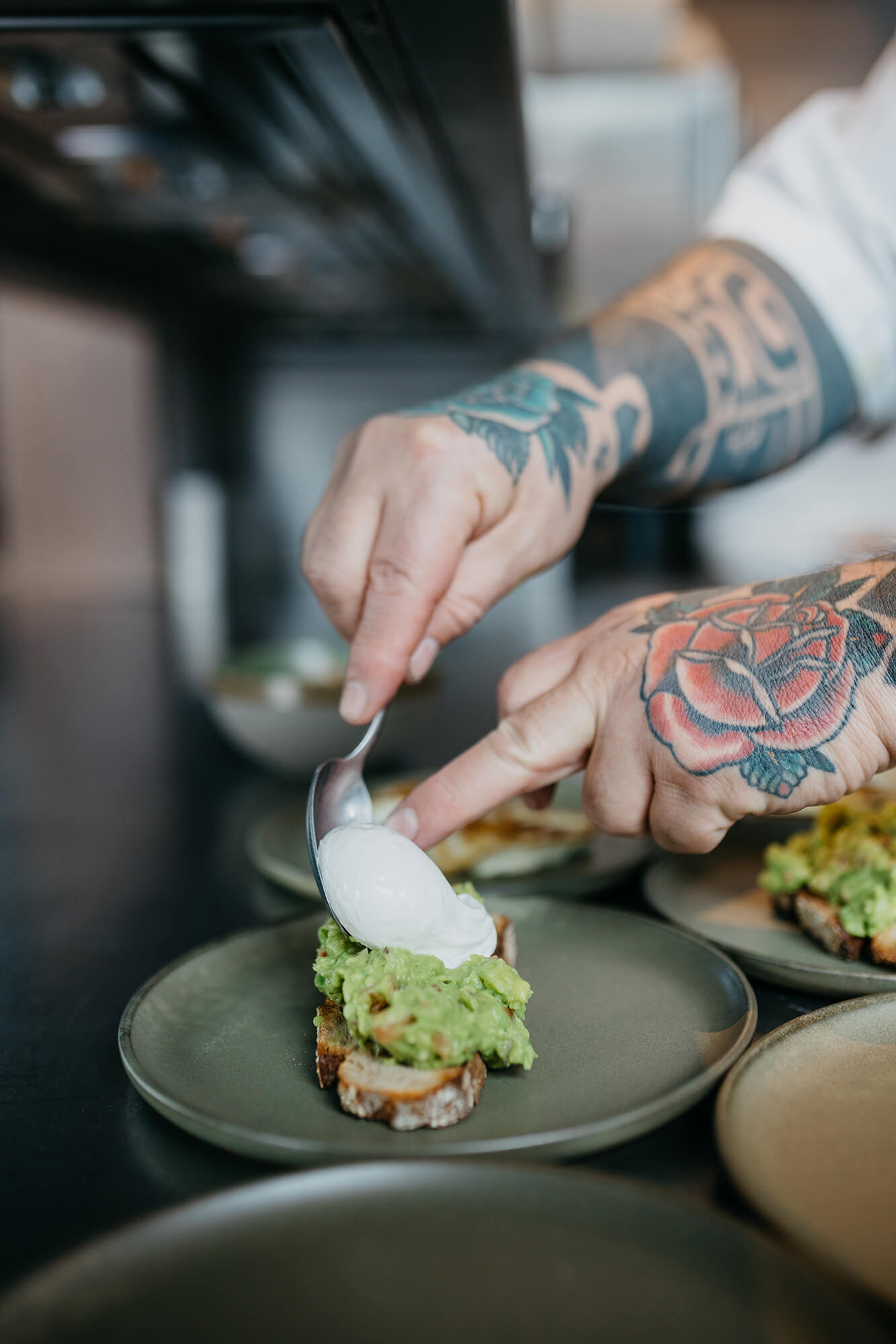 Close-up of a chef's tattooed hands spooning an egg over avocado toast at Vessel's kitchen