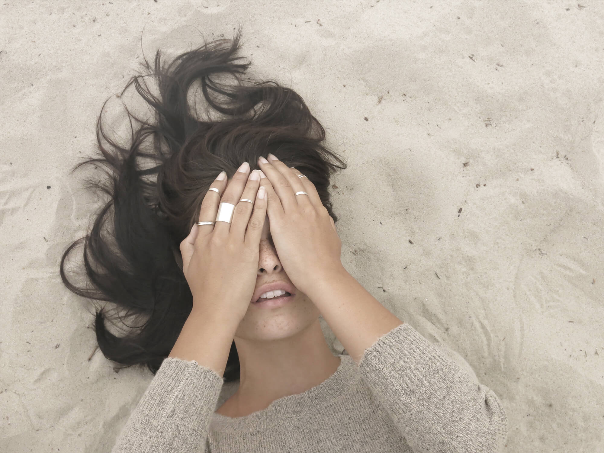 Overhead shot of a woman lying in the sand covering her eyes with both ringed hands, dark hair fanned out around her