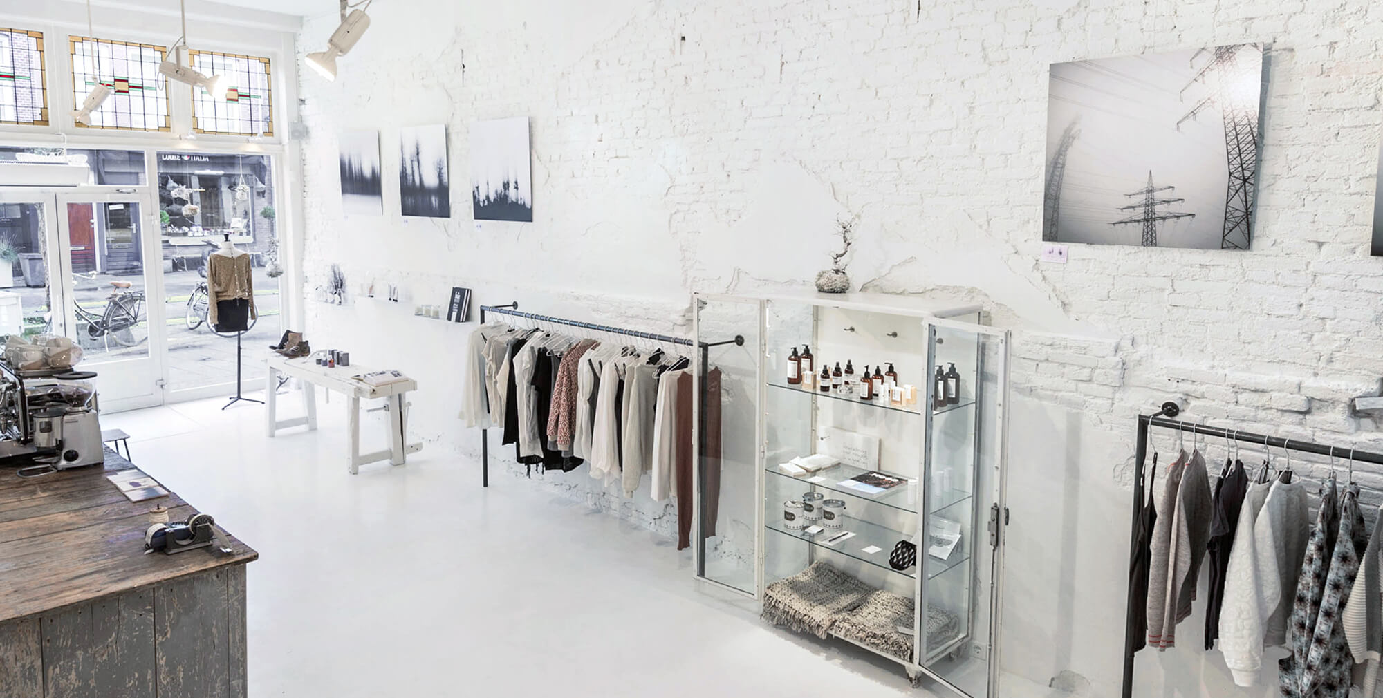 Wide view of the COTTONCAKE retail interior with whitewashed brick walls, framed photography, a vintage coffee counter, clothing racks, and a glass display cabinet of products